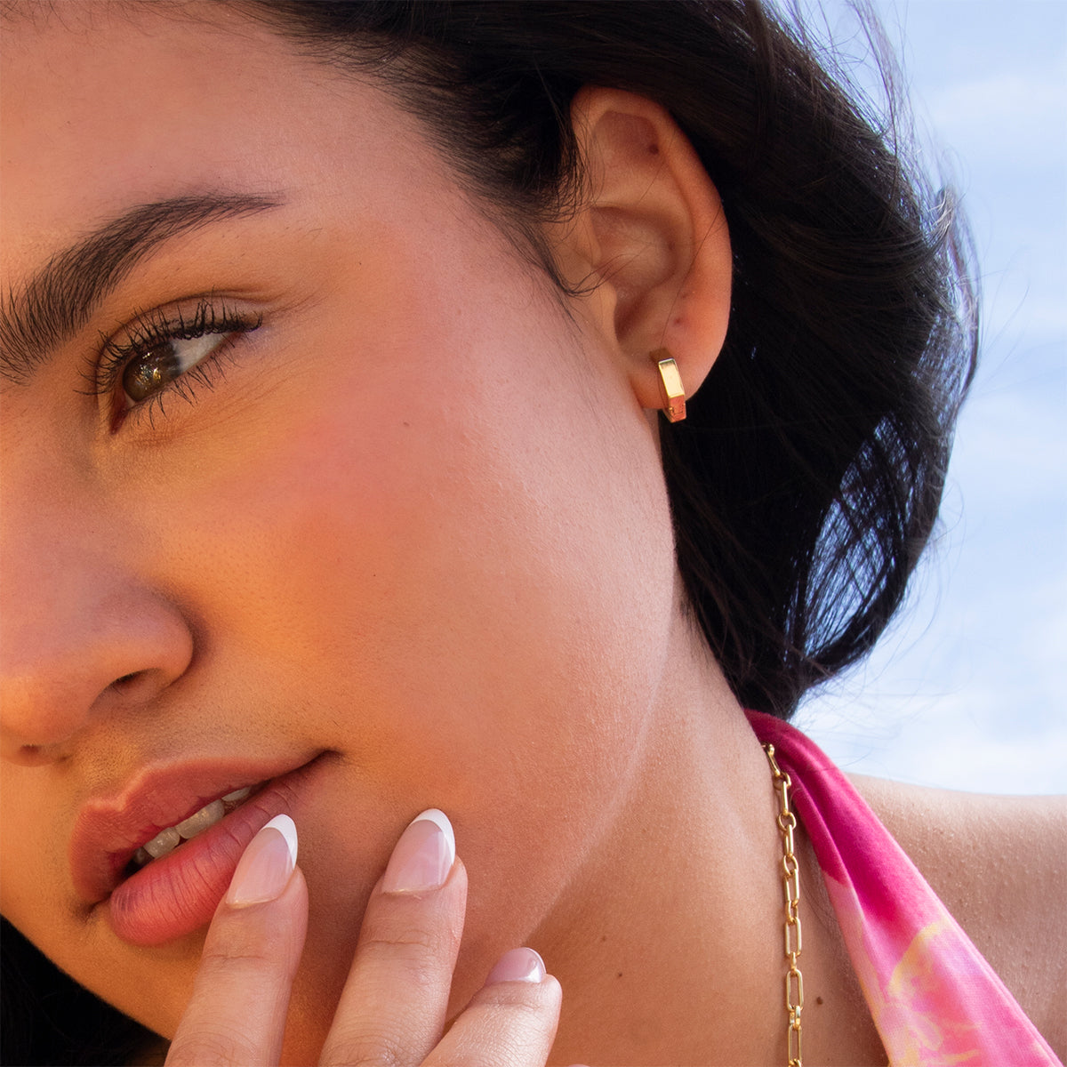 A close-up of a woman wearing the Ari Hoops gold earrings, with glossy lips, manicured nails, wavy dark hair, and a pink halter top. She touches her face against a backdrop of blue sky.