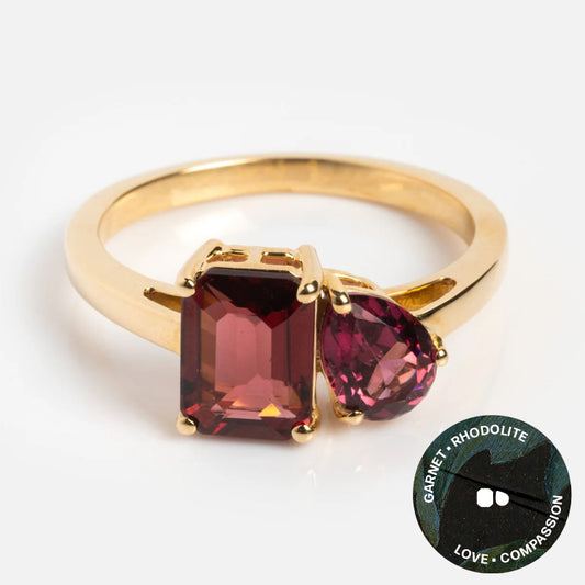 The Solid Gold Toi et Moi Colorful Gemstone Ring showcases two garnet stones—one rectangular, one teardrop—set side by side. An inset circle reads Garnet · Rhodolite · Love · Compassion. A striking, unique gemstone jewelry piece.