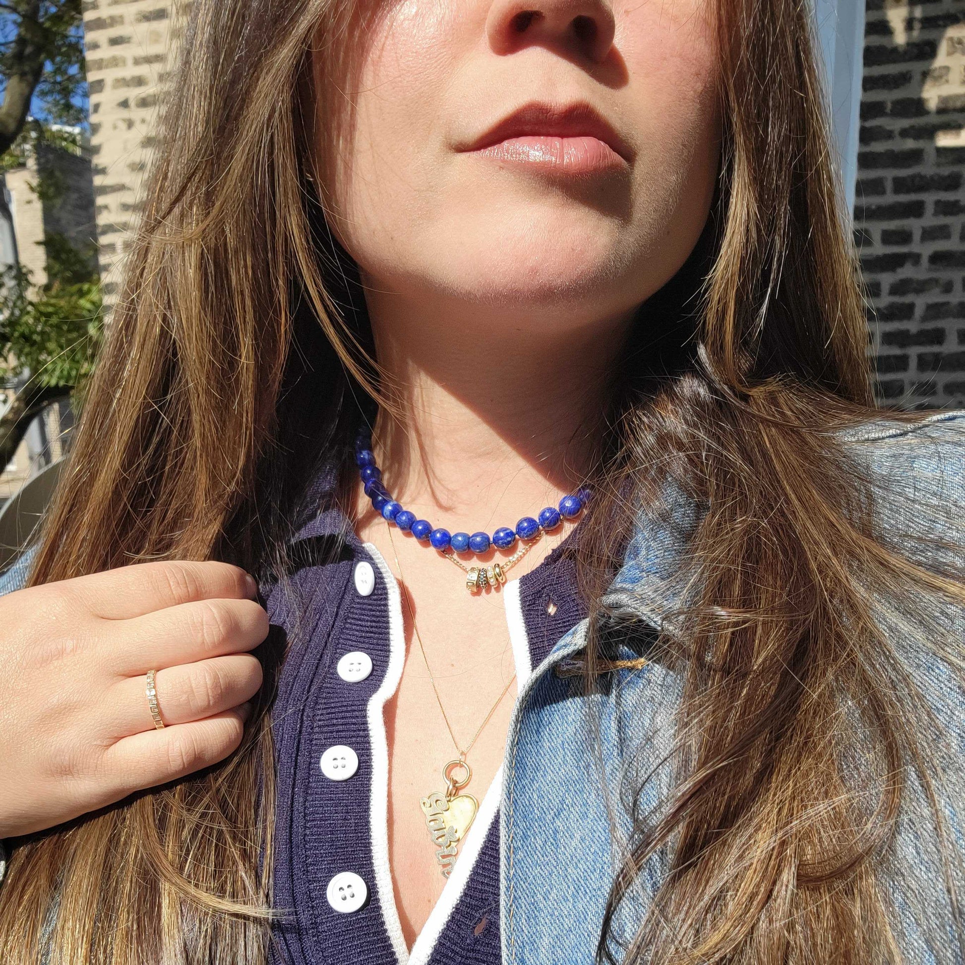 A woman with long brown hair wears the Sale! Beaded Gemstone Necklace, paired with a navy collared top and denim jacket, standing outside in sunlight against a brick wall.
