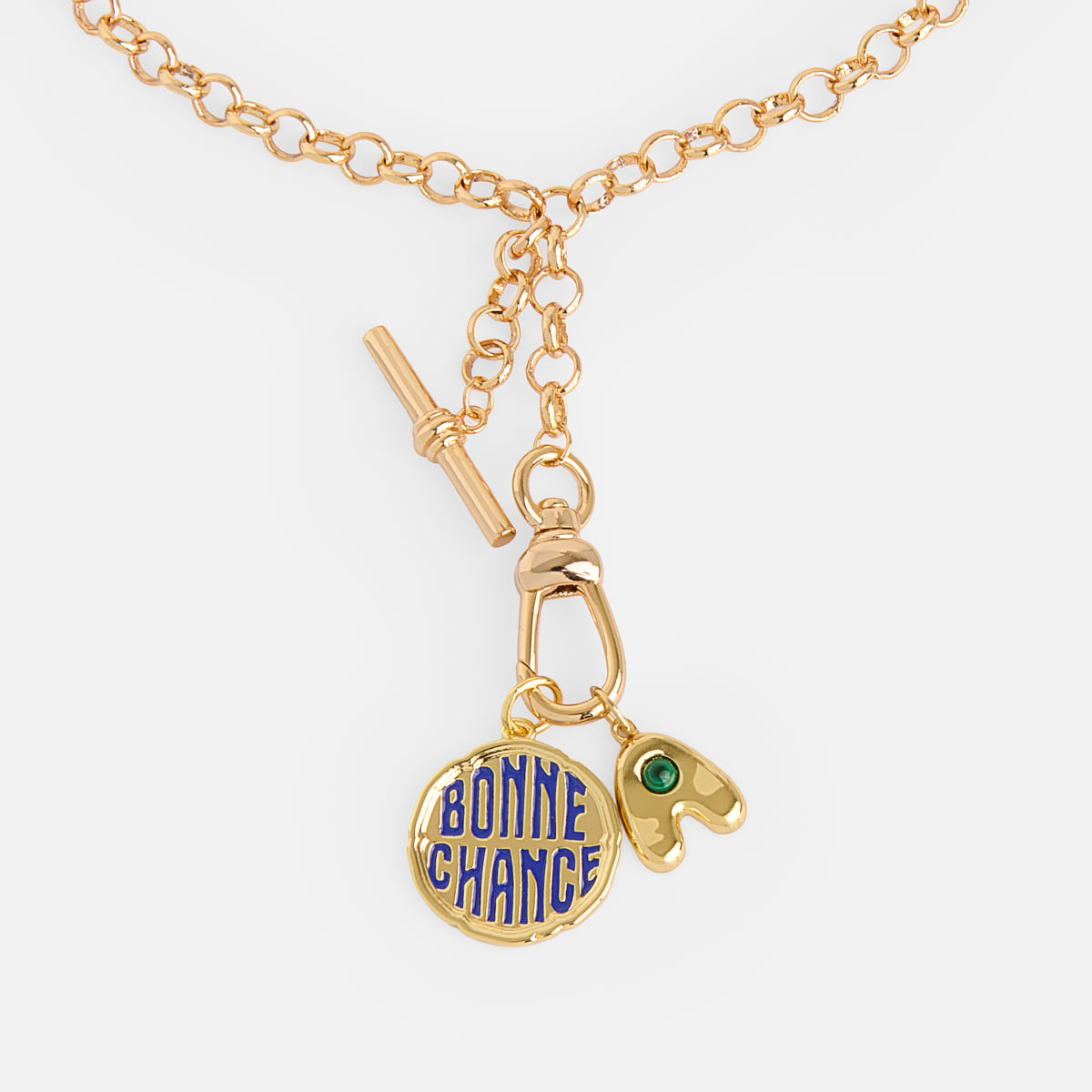 The Cameron Charm Holder Necklace is a gold chain with a toggle clasp, featuring a blue-lettered BONNE CHANCE round charm and an irregular gold charm accented by a small green gem.