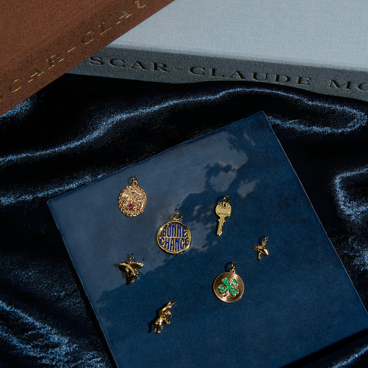 Six gold Charms by Mod and Jo—a key, "QUIET CHANCE" coin, four-leaf clover, star, bird, and bear—are displayed on a blue ceramic tile with books and dark velvet fabric in the background.