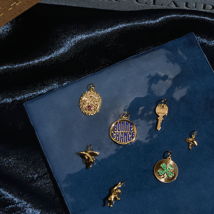 Seven Charms by Mod + Jo, including a key, clover, airplane, bunny, bird, oval, and Bonne Chance pendant, are arranged on a glossy dark blue surface with a dark cloth and book in the background.