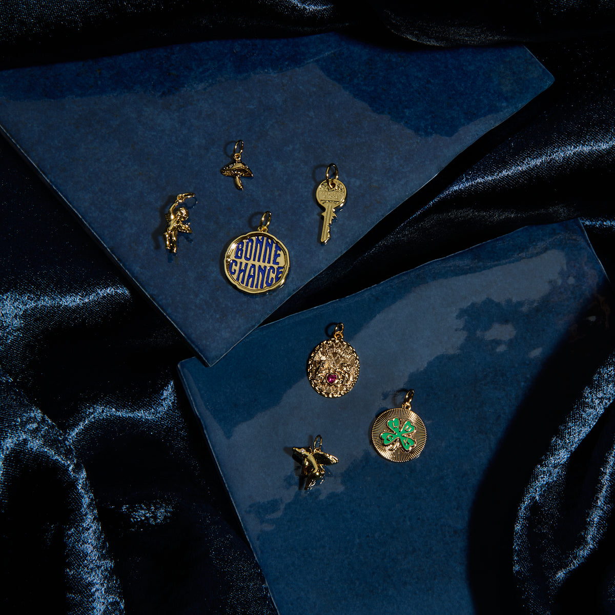Nine gold Charms by Mod and Jo, featuring a key, ballerina, Bonne Chance disc, four-leaf clover, and other pendants, are displayed on blue textured surfaces with dark velvet in the background.