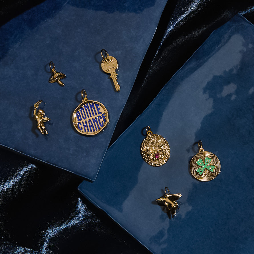 Charms by Mod + Jo features gold plated charms—a hummingbird, key, astronaut, blue enamel "Bonne Chance" pendant, coin, four-leaf clover, and bird—arranged on glossy blue and black surfaces.