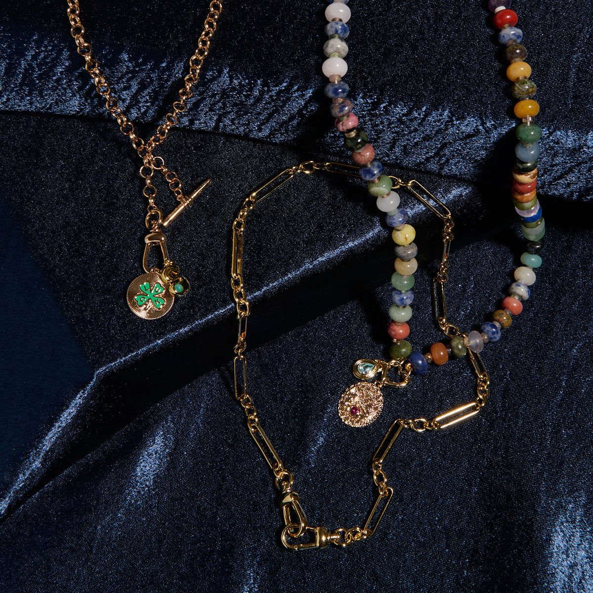 Three necklaces rest on dark blue velvet: the Liam Watch Chain Charm Holder Necklace, a gold chain with a round gem pendant, and a beaded necklace with multicolored stones.