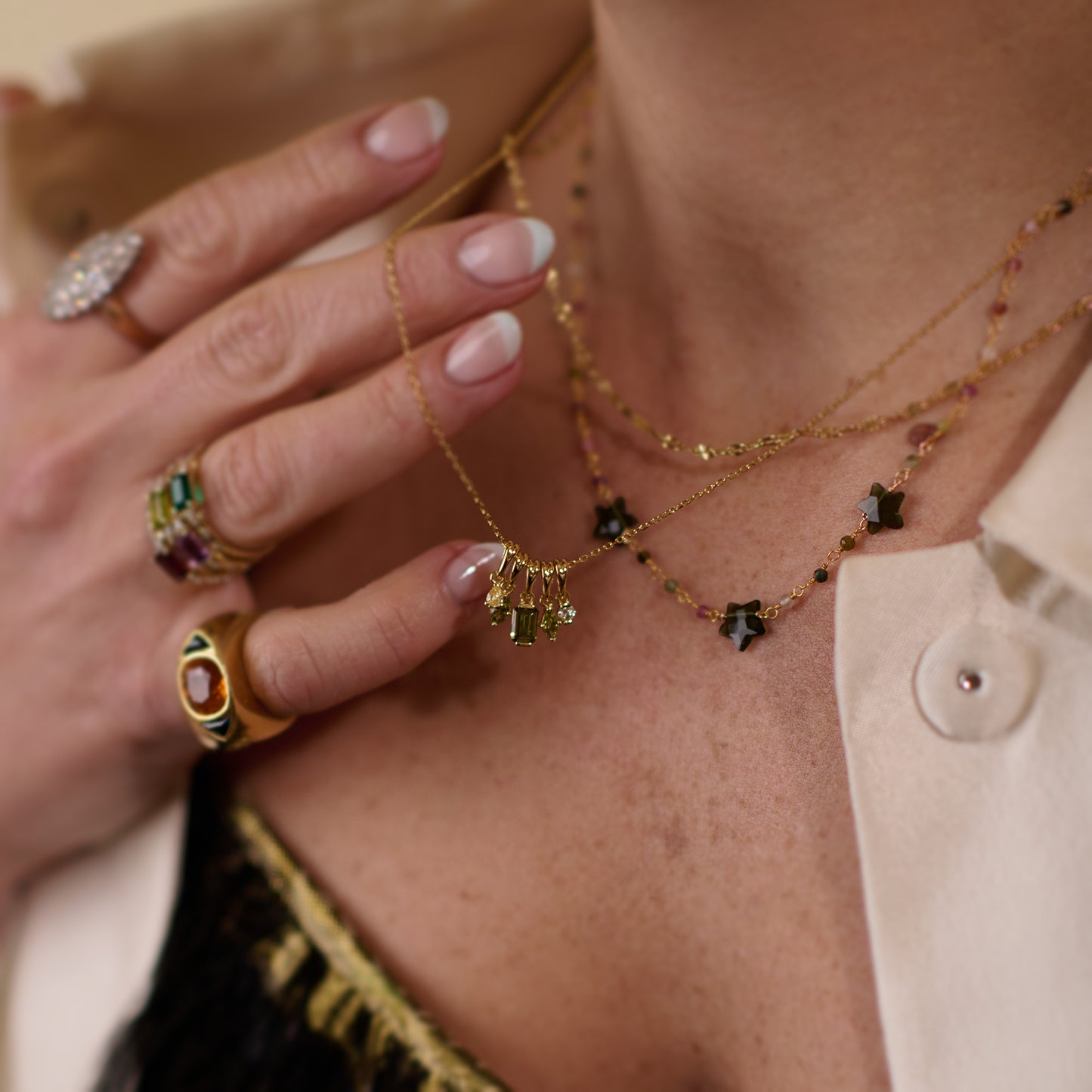 A close-up of a woman’s hand touches layered gold plated necklaces, featuring the Collier Bangalore Necklace with green tourmaline pendants. She wears gemstone rings and a cream shirt over a black lace top, with naturally manicured nails.