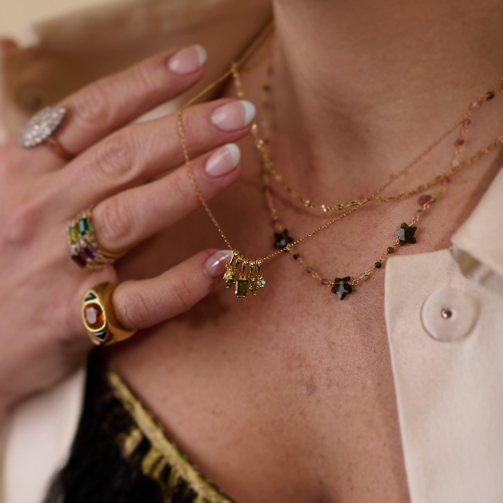 A close-up of a woman’s hand touches layered gold plated necklaces, featuring the Collier Bangalore Necklace with green tourmaline pendants. She wears gemstone rings and a cream shirt over a black lace top, with naturally manicured nails.