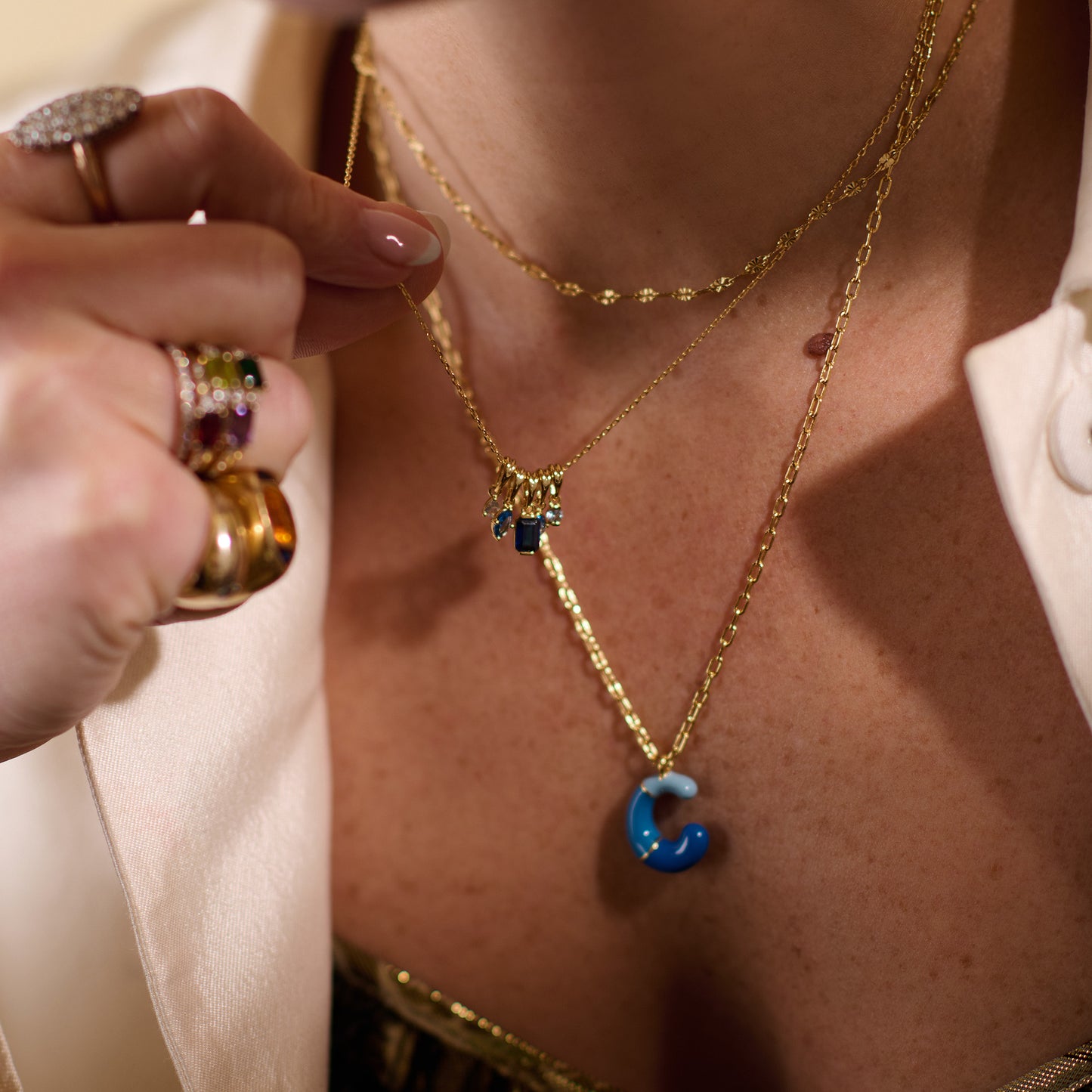 A close-up of a person wearing layered gold necklaces, featuring the Shades of You Ombre Birthstone Charm Necklace with a blue "C" pendant. The look is completed with gold rings and an off-white blazer.