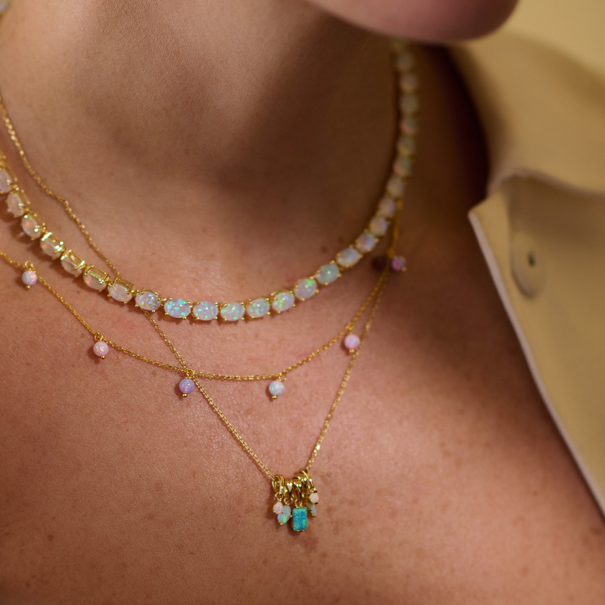 A close-up of a person wearing the Baby Blue Opal Eternal Tennis Necklace layered with gold-plated necklaces featuring pastel gemstone accents, shown against light skin and a beige collared shirt.