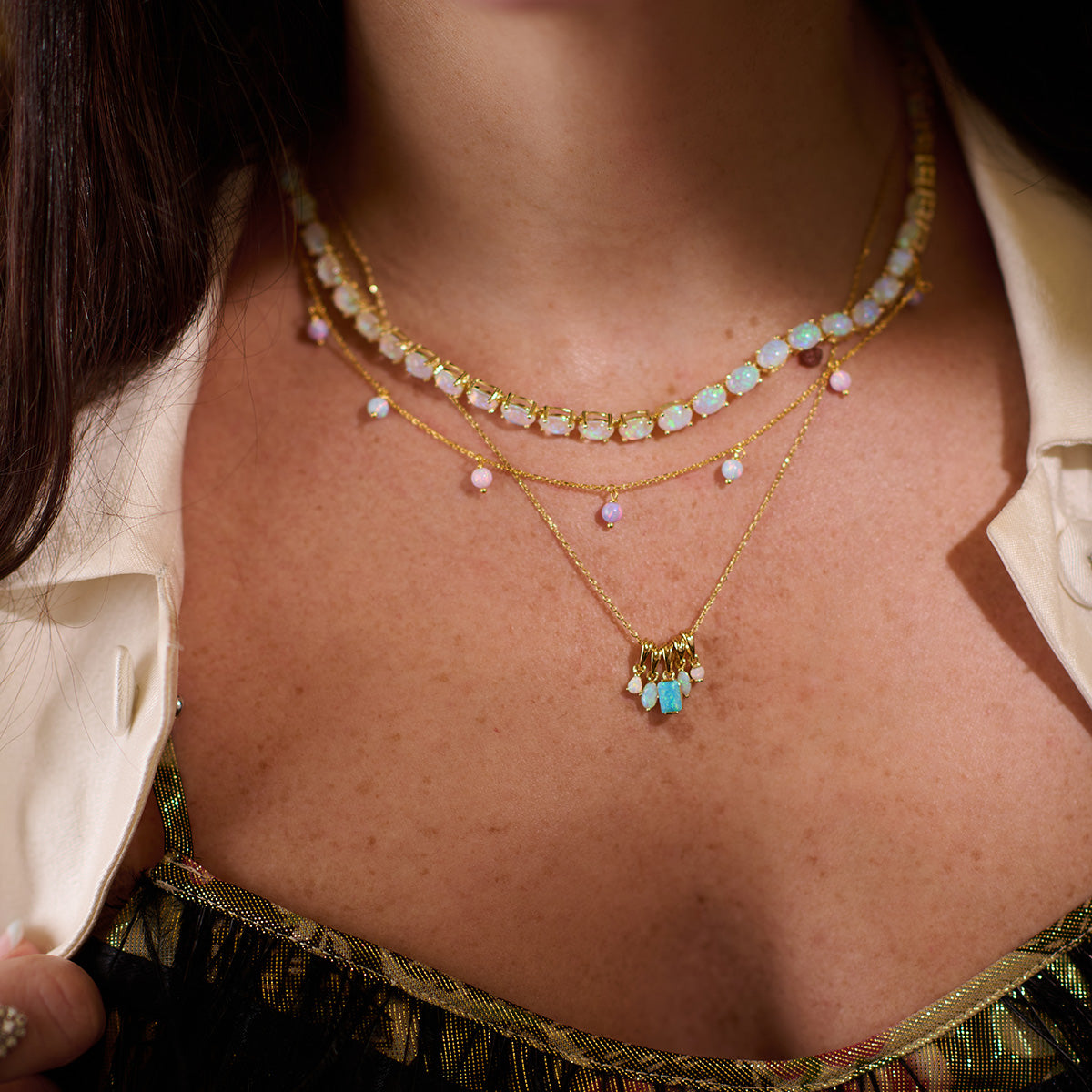A woman wearing the Baby Blue Opal Eternal Tennis Necklace and a gold plated chain with colorful gemstones, dressed in a patterned top and off-white shirt, shown from shoulders to just below the collarbone.