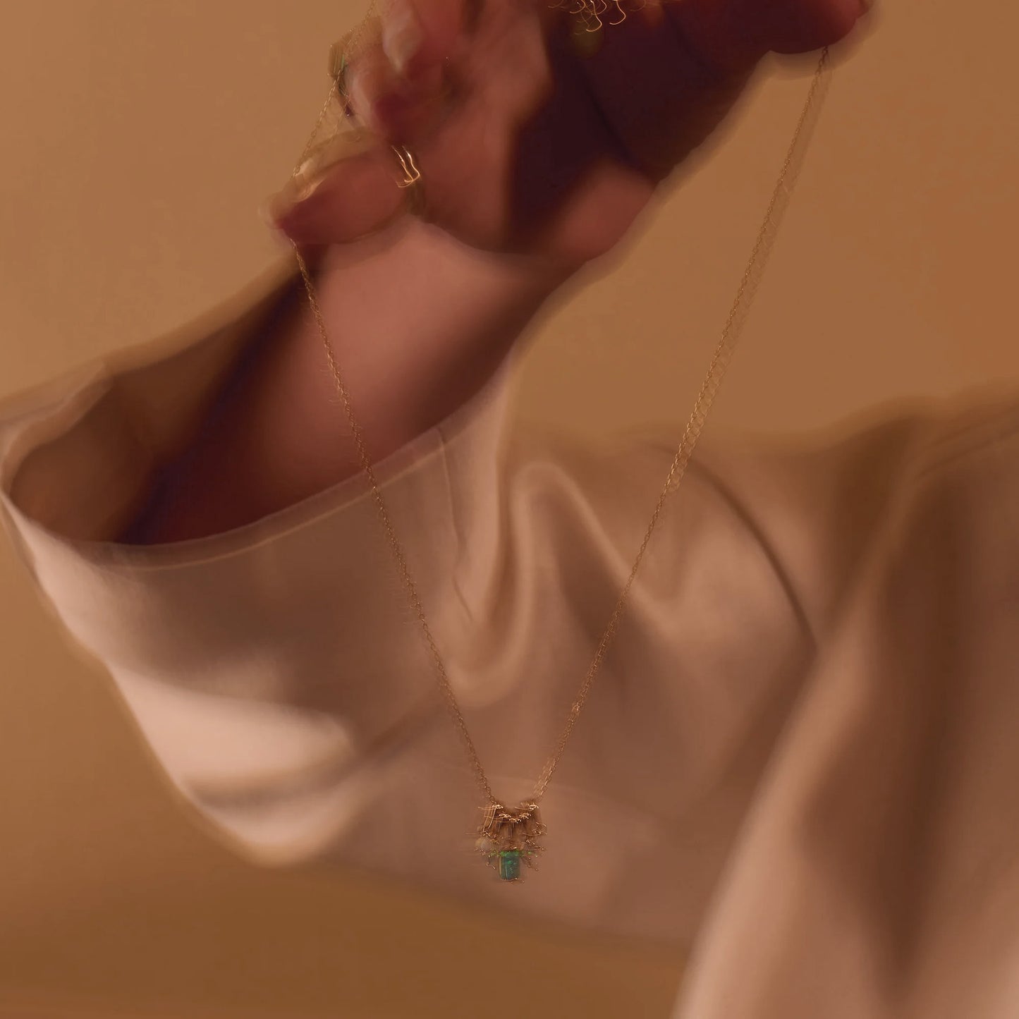 A hand holds up the Shades of You Ombre Birthstone Charm Necklace, featuring a delicate gold chain with a small pendant, against a softly blurred warm background. The wearer sports a light-colored, long-sleeved garment. A Local Eclectic exclusive.