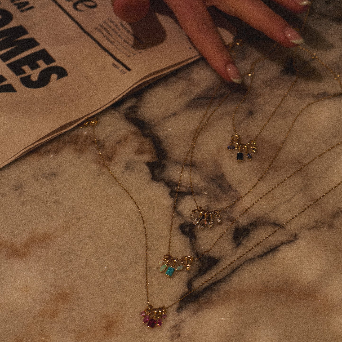 A hand with manicured nails rests on a marble surface next to a newspaper. Several gold necklaces, including the Shades of You Ombre Birthstone Charm Necklace, are displayed on the marble.
