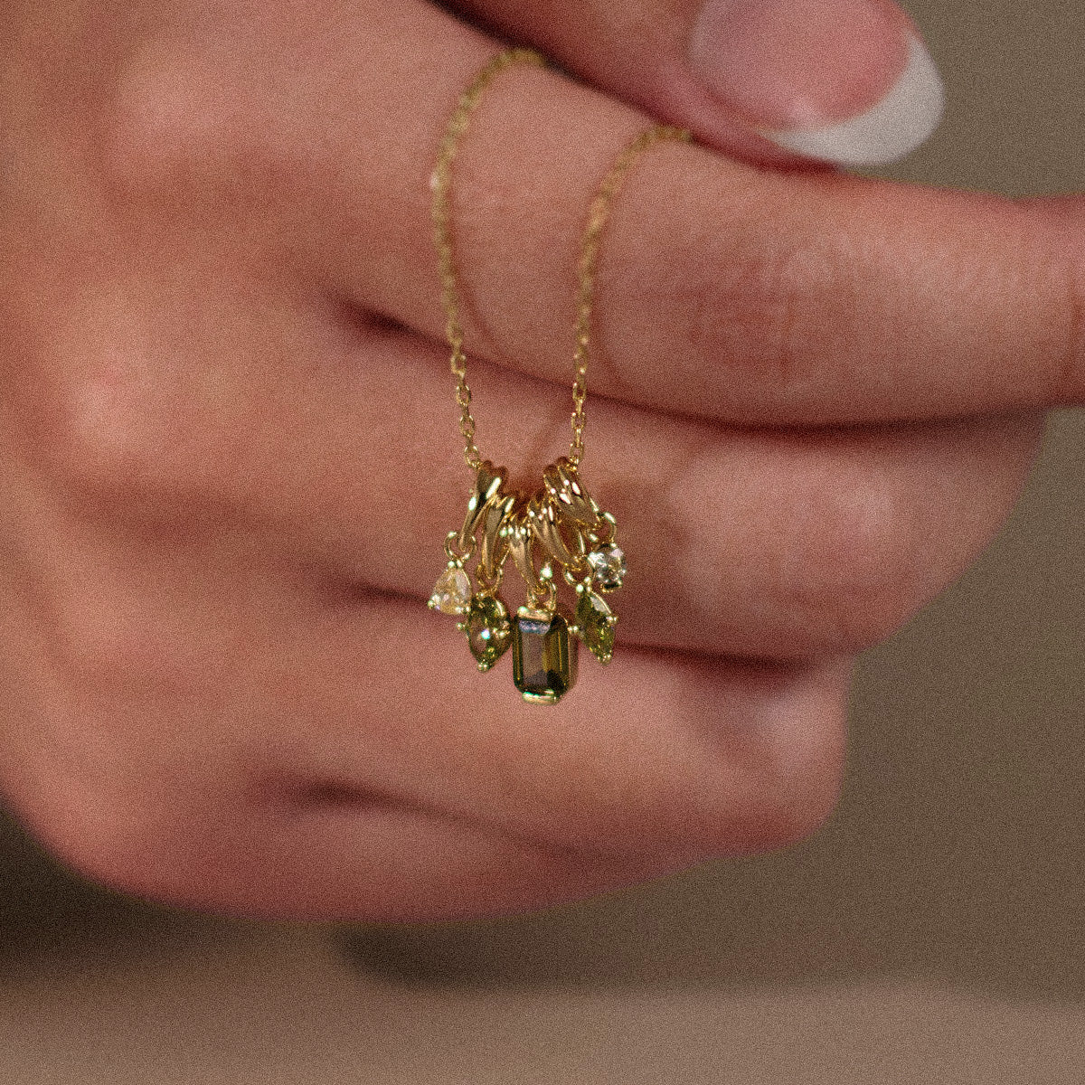 A hand with a French manicure holds the Shades of You Ombre Birthstone Charm Necklace, a delicate 14k gold plated piece featuring several gold and gemstone charms, including a rectangular green stone and teardrop-shaped gems.