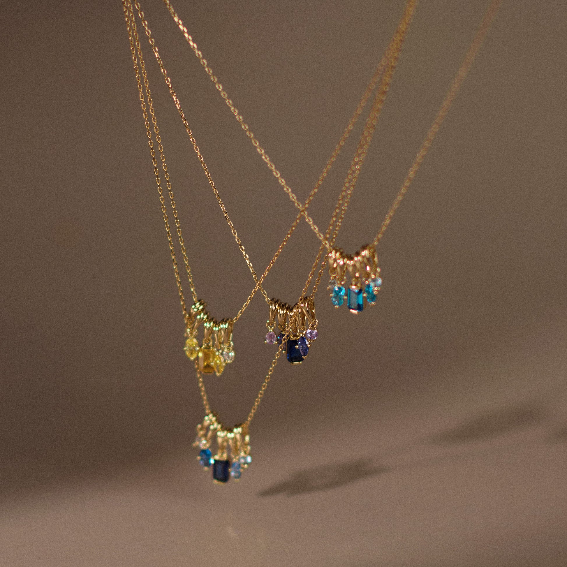 Three Shades of You Ombre Birthstone Charm Necklaces hang side by side, each with small, colorful blue, yellow, and purple ombre pendants, displayed against a softly lit neutral background.
