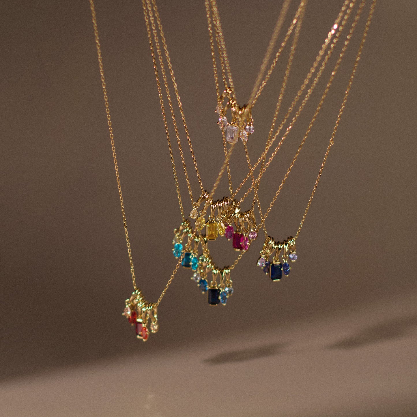 The Shades of You Ombre Birthstone Charm Necklaces, featuring colorful gemstone pendants in various shapes, hang closely together against a blurred neutral background, creating a vibrant and elegant birthstone display.