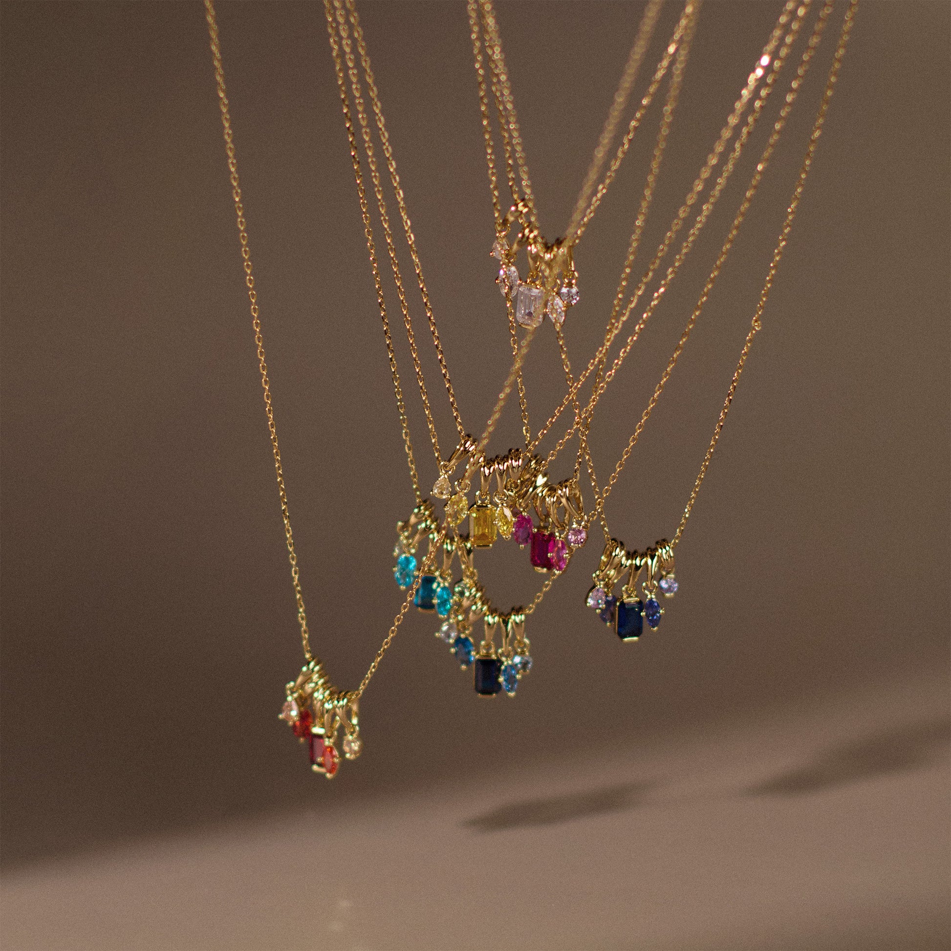 The Shades of You Ombre Birthstone Charm Necklaces, featuring colorful gemstone pendants in various shapes, hang closely together against a blurred neutral background, creating a vibrant and elegant birthstone display.