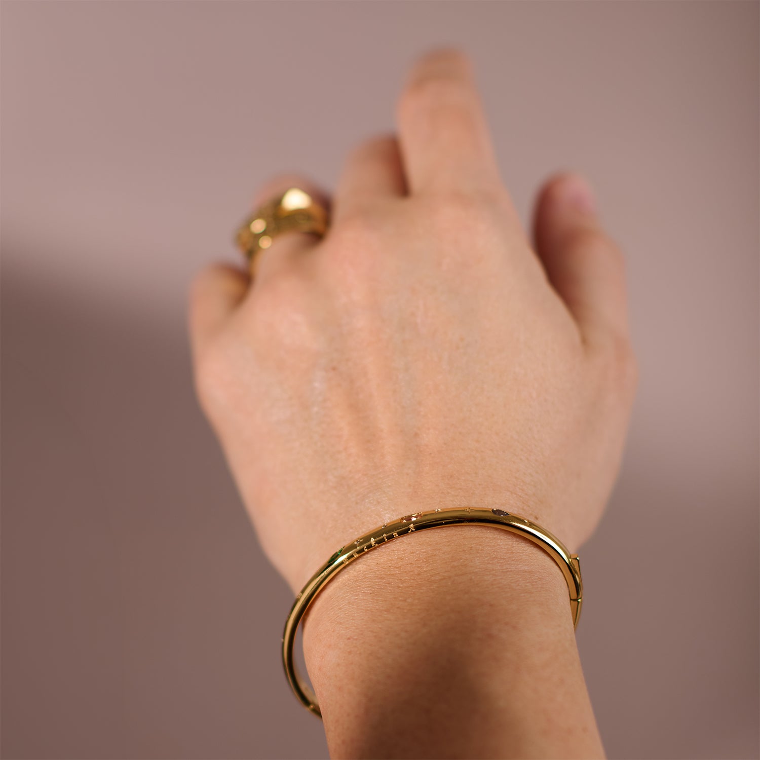 A hand wears the Hidden Curse Word Fuck Off Bracelet—a simple gold bangle—along with a gold ring, set against a softly blurred light brown background.
