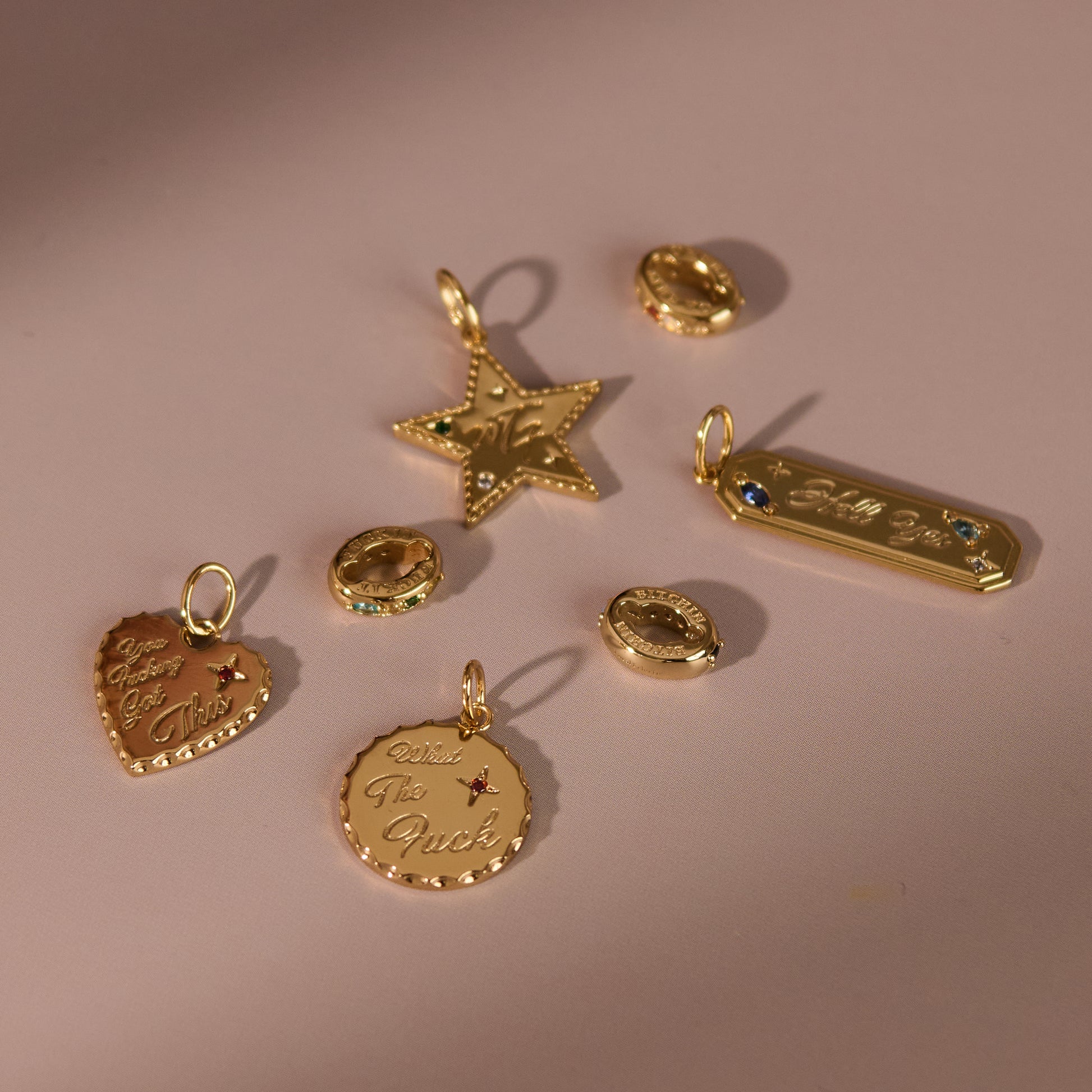 The Curse Word Charm, available in shapes like star, heart, circle, and bar, is engraved with text and adorned with colorful gemstones, all displayed on a soft pink surface.