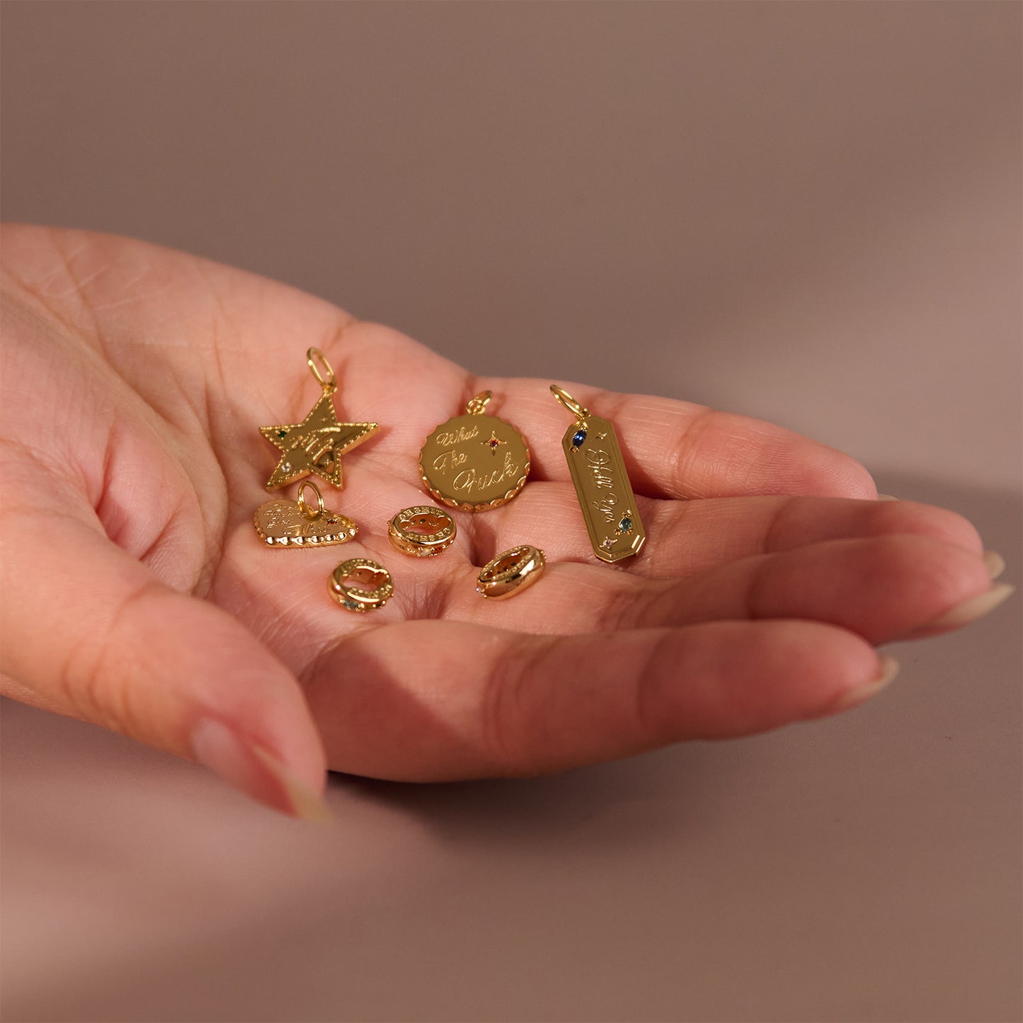 A hand holds several small gold charms, including the Hidden Curse Word Spacer Charm, a star, a heart, two rings, an engraved round disc, and a rectangular pendant with a tiny blue gem against a soft, neutral background.