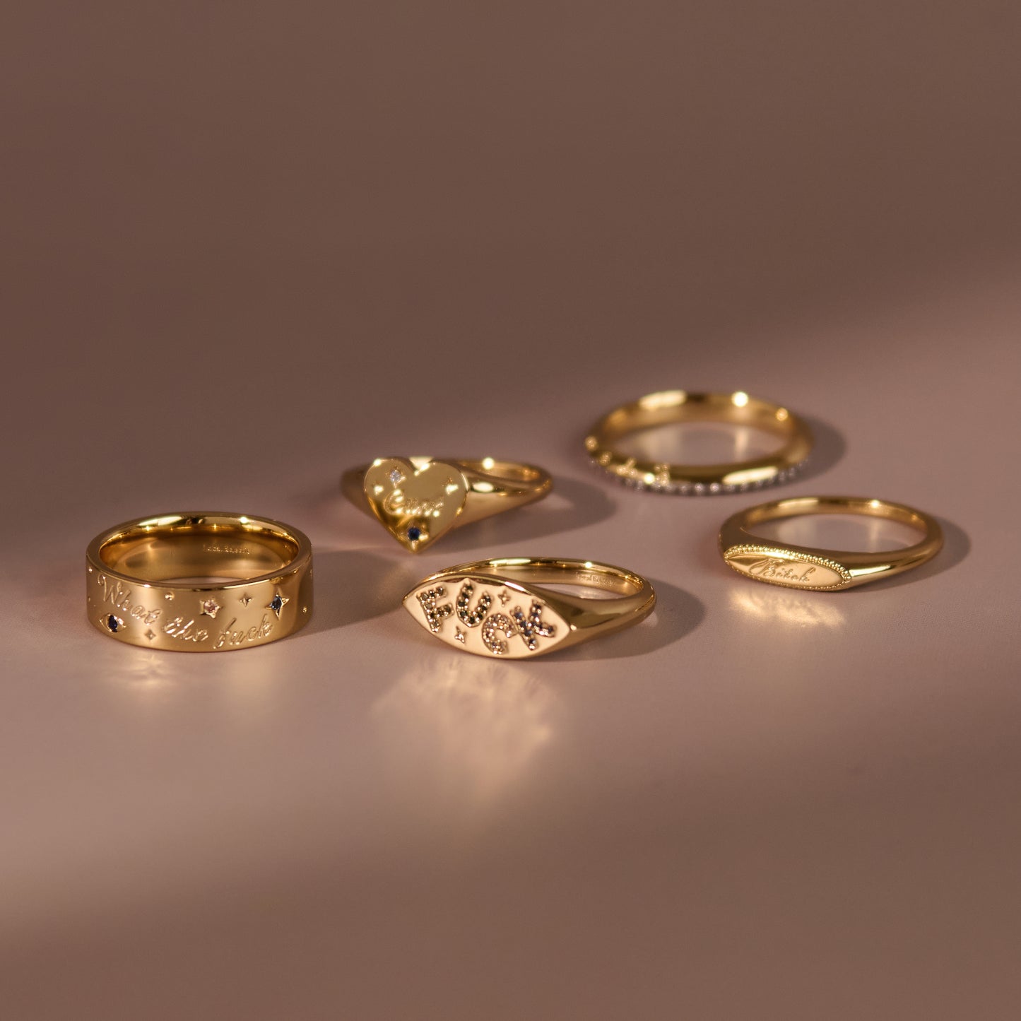 Five Colorful Language Fuck Signet Rings with engraved and textured designs are displayed on a softly lit surface. Their arrangement casts gentle shadows, emphasizing their metallic shine and intricate details.
