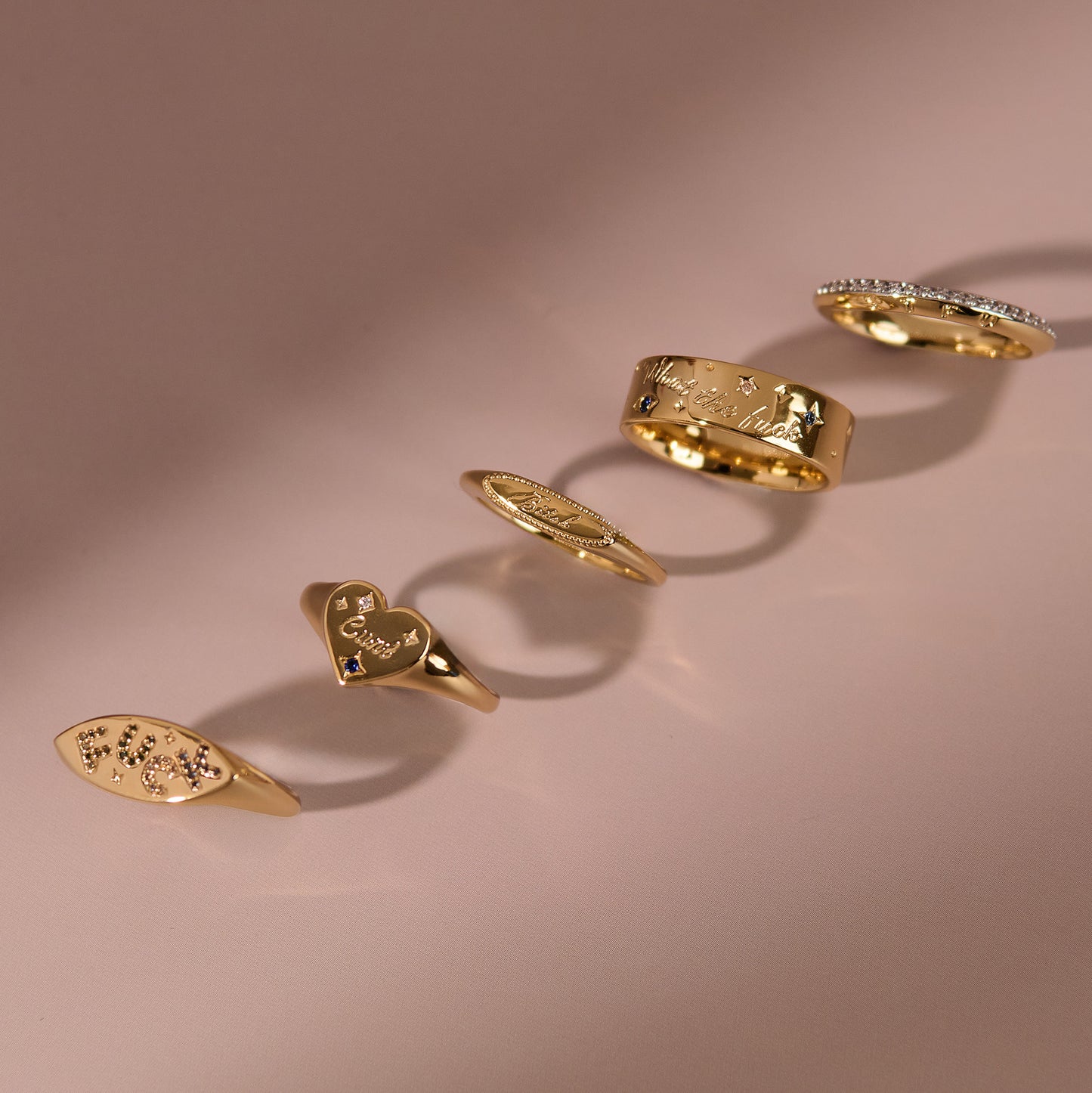 The Colorful Language Fuck Signet Ring, 14k gold plated with an engraved design, is displayed on a soft pink surface, casting gentle shadows and featuring unique embossed patterns for a bold statement.