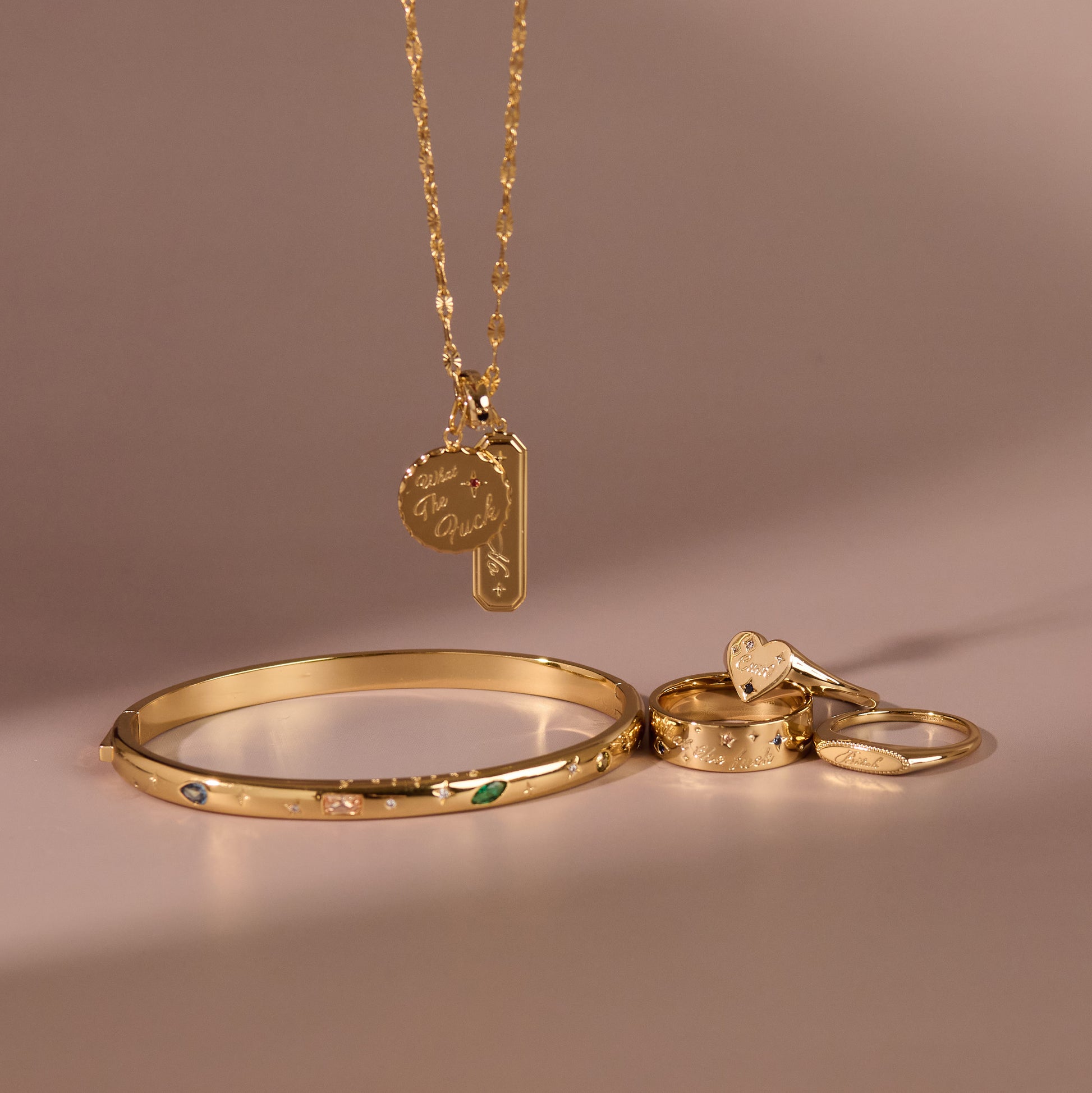 A gold jewelry set featuring a bracelet with colorful stones, two rings, and a necklace adorned with two engraved Curse Word Charm gold pendants, all displayed on a neutral background.
