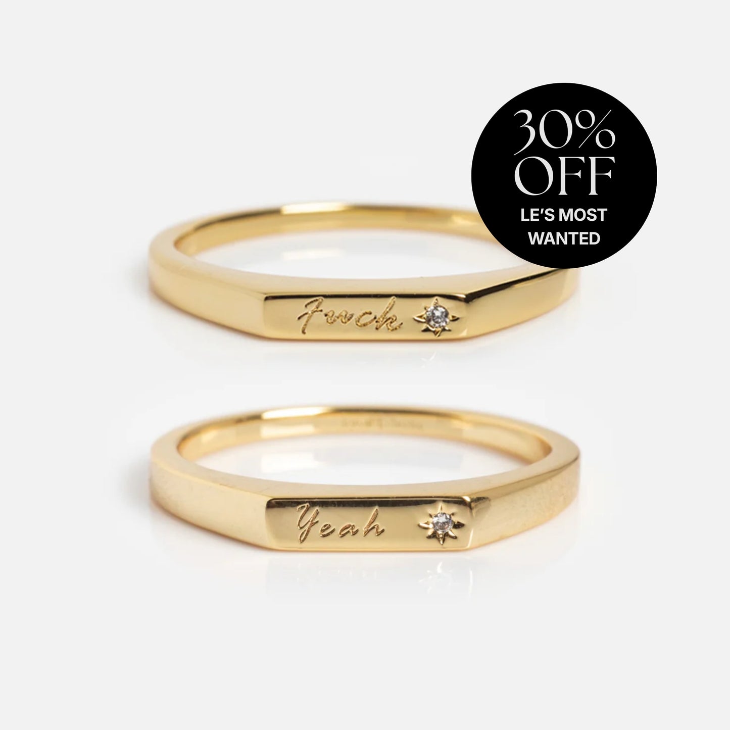 Two Tiny Conversation Stacking Rings in 14k yellow gold plate, each engraved with "F—ck" and "Yeah" and set with a sparkling white CZ, are shown on a white background. A black circle top right says “30% OFF LES MOST WANTED.”.