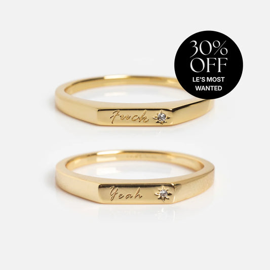 Two Tiny Conversation Stacking Rings in 14k yellow gold plate, each engraved with "F—ck" and "Yeah" and set with a sparkling white CZ, are shown on a white background. A black circle top right says “30% OFF LES MOST WANTED.”.