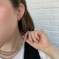Close-up of a person wearing Classic Gem Huggie Hoops (14k gold plated) and holding a twisted gold-and-silver chain necklace, with peach-colored nail polish, against a white brick wall.