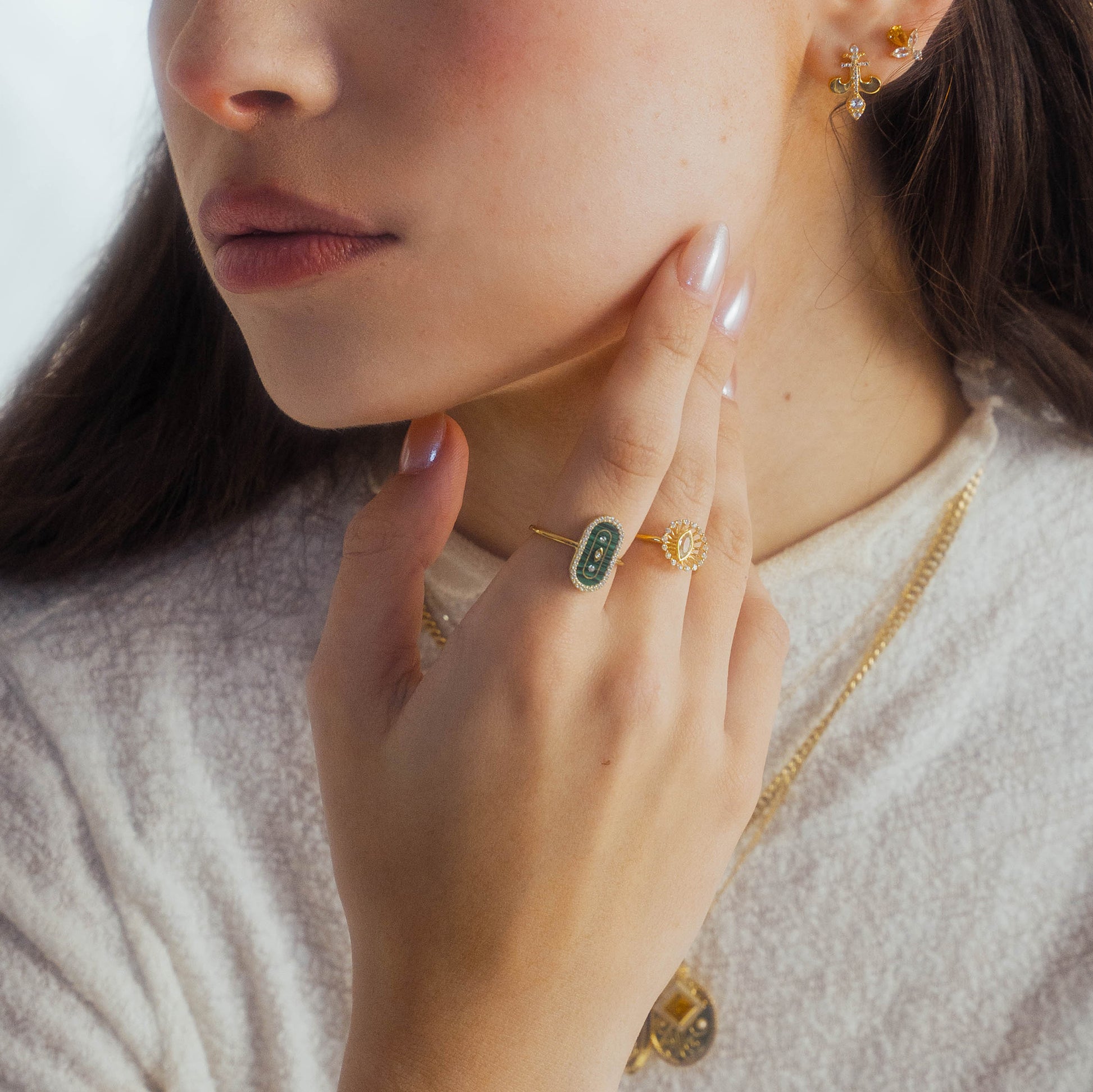 A woman with long brown hair touches her chin, wearing a white top, 14k yellow gold vermeil earrings, layered gold necklaces, and the So Art Deco Cocktail Ring with vibrant Malachite. Only her lower face and hand are visible.