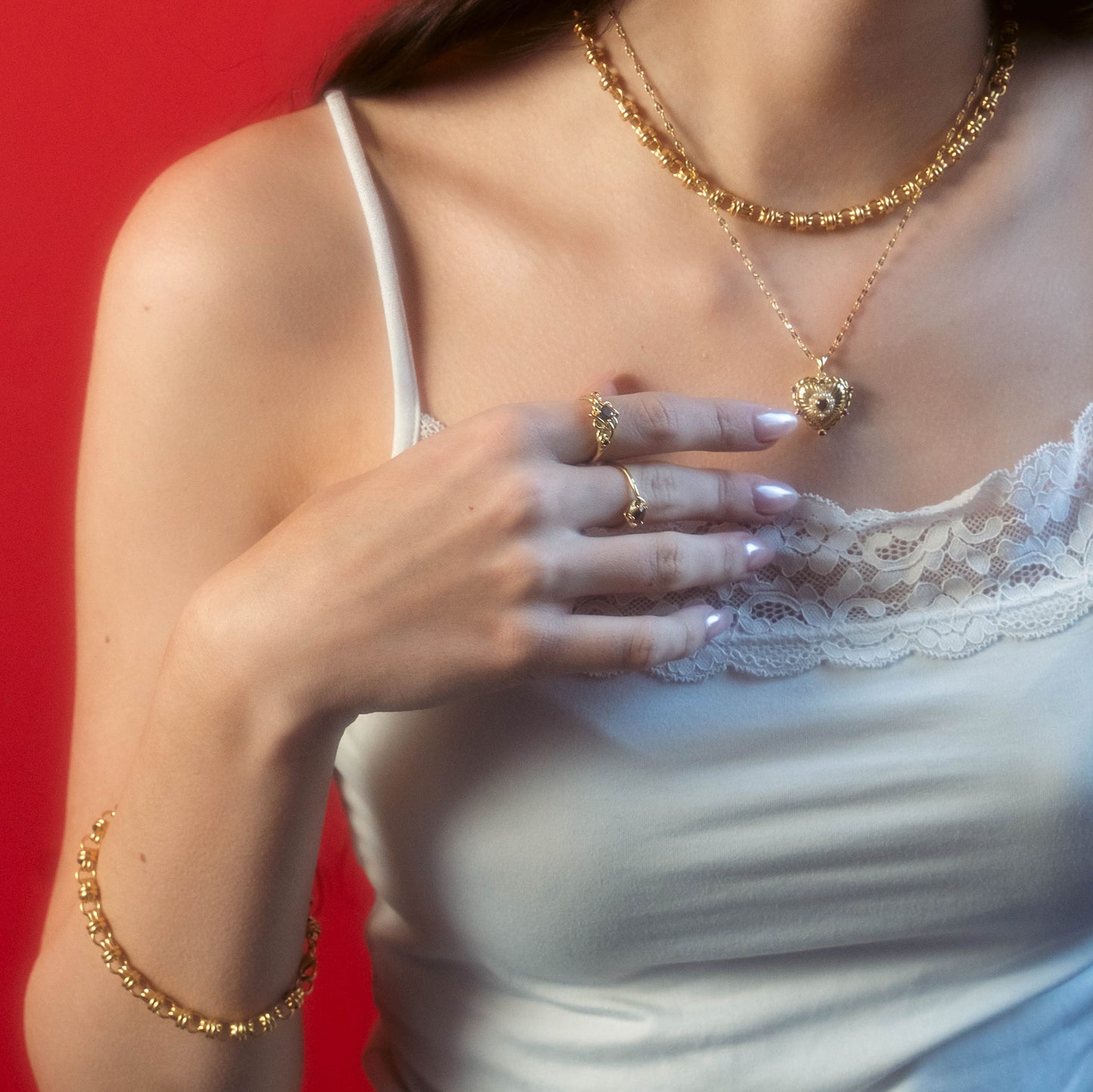 A woman with light-manicured nails wears a white lace camisole and displays gold jewelry, featuring the Lover's Knot Heart Ring, layered necklaces, bracelets, and rings against a red background.