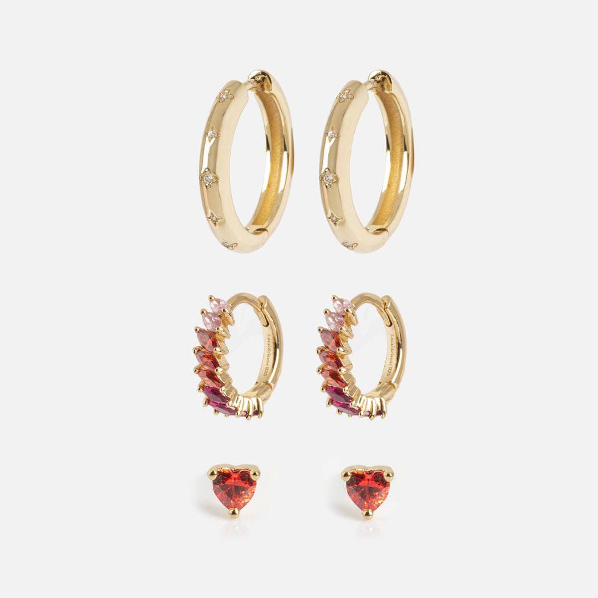 The Bold Birthstone Earring Set features three gold pairs: diamond-accented hoops, pink and red gemstone hoops, and heart-shaped red gemstone studs, all shown vertically against a white background.