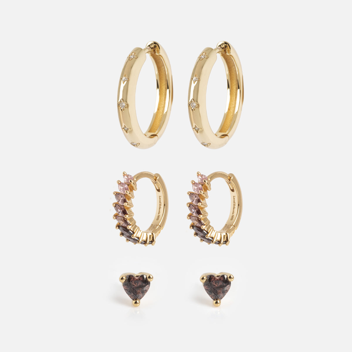 The Bold Birthstone Earring Set features three pairs: gold hoop earrings with small diamonds, smaller hoops with pink stones, and heart-shaped studs with dark gemstones, all displayed on a white background.