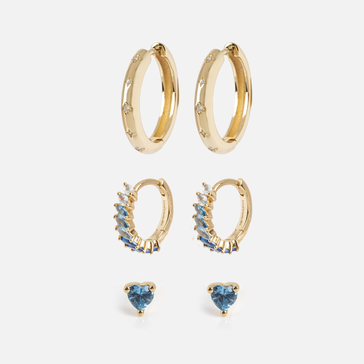 The Bold Birthstone Earring Set includes three gold pairs: plain hoops with small gems, smaller hoops lined with blue and white stones, and stud earrings with blue heart-shaped stones, all shown on a white background.