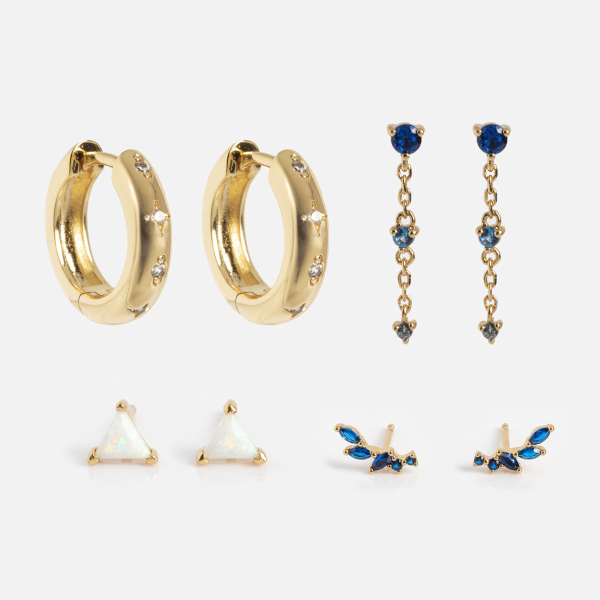 The Bold Birthstone Earring Set includes four pairs: gold hoops with small gems, gold drop earrings with blue stones, gold triangle studs with white centers, and wing-shaped studs adorned with blue and clear stones.