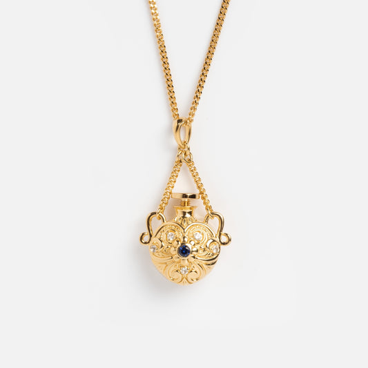 The Juliet Vial Pendant Necklace features an ornate gold flask-shaped pendant with intricate scrollwork and a central blue sapphire, hanging from a delicate gold chain—a stunning piece of vintage-inspired jewelry.