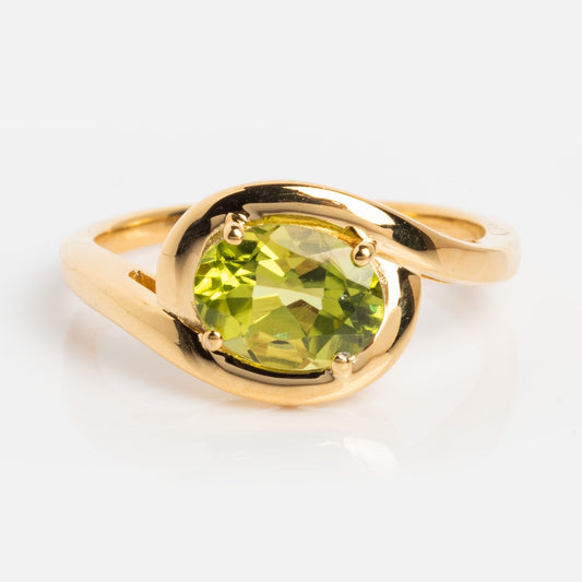 The Peridot Poetry Statement Ring is a vintage-inspired 14k gold vermeil ring with a round, faceted green peridot held by four prongs and a gracefully twisting band, shown against a plain white background.