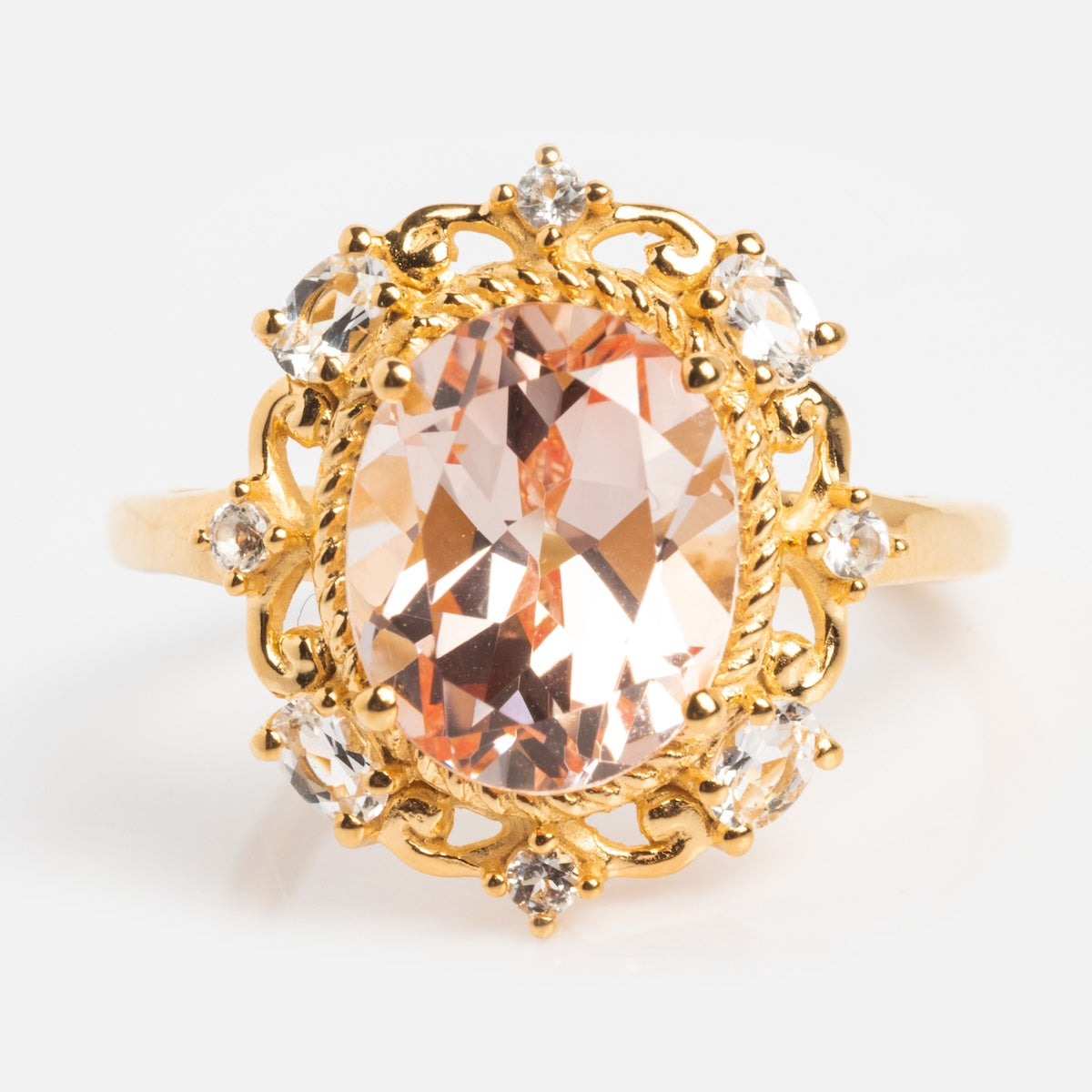 The Royal Blush Cocktail Ring is a 14k gold vermeil ring with a large oval peach gemstone, accented by small clear stones and intricate gold detailing, set on a plain white background.
