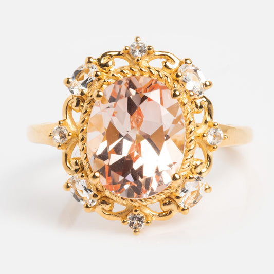 The Royal Blush Cocktail Ring is a 14k gold vermeil ring with a large oval peach gemstone, accented by small clear stones and intricate gold detailing, set on a plain white background.