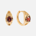 The Gilded Garnet Huggie Hoops are gold vermeil earrings with oval red gemstones at the center, encircled by sparkling white topaz, shown on a plain white background.