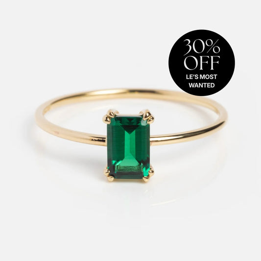 A slim gold ring features a baguette-cut green birthstone at its center. A black circle in the corner displays: 30% OFF LES MOST WANTED. This is the Solid Gold Essential Baguette Birthstone Ring.