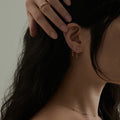 A woman with dark hair touches her head, displaying Solid Gold White Topaz Dangle Chain Huggies. Only the side of her face and upper neck are visible against a neutral background.