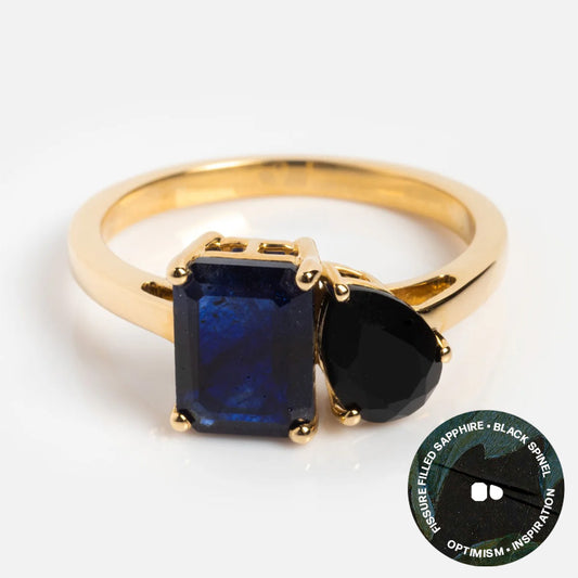 The Solid Gold Toi et Moi Colorful Gemstone Ring features a rectangular blue sapphire and a teardrop black spinel, labeled “Pressure filled sapphire + black spinel, optimism + inspiration.” Perfect for unique gemstone jewelry lovers.