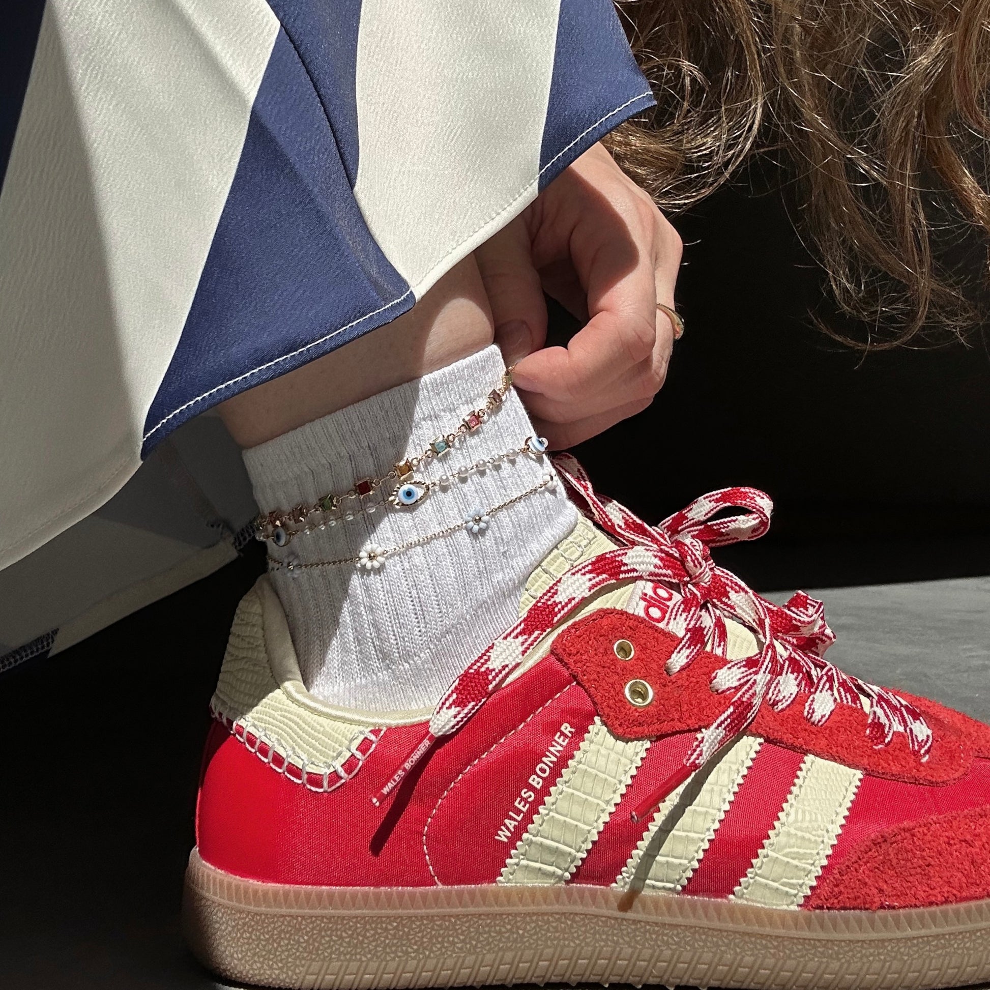 A person wearing red and white Adidas sneakers, white socks, blue-and-white pants, and a gold plated Evil Eye Anklet featuring a delicate evil eye charm is shown.