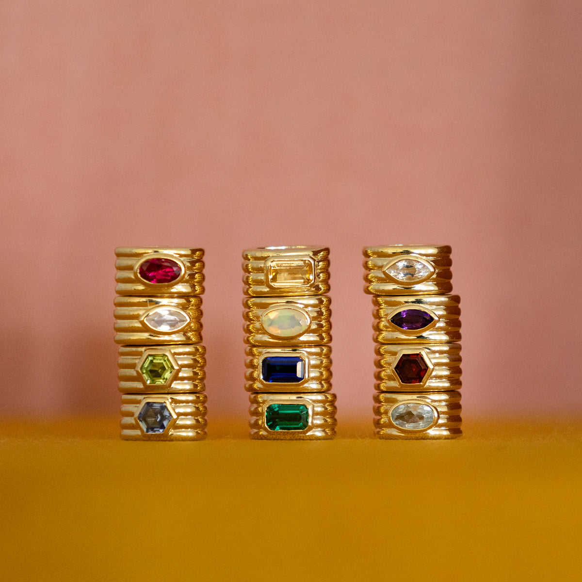 Three stacks of Solid Gold Bold Birthstone Spacer Charms, each set with colorful gemstones in various shapes, displayed upright against a blurred peach and yellow background.