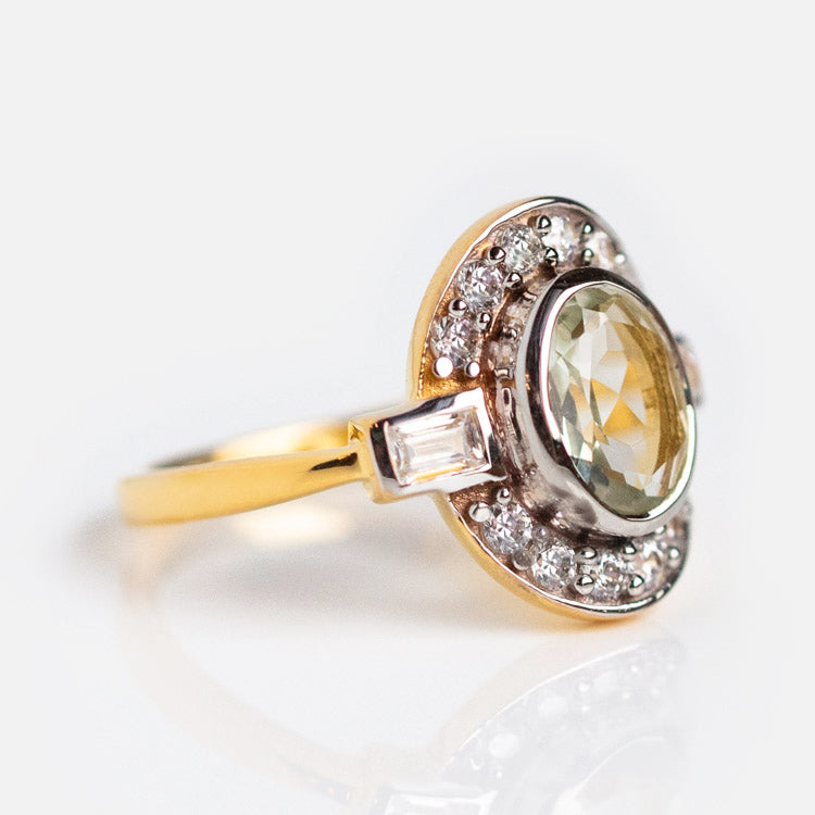 The Oval Deco Green Amethyst and CZ Ring features an oval, faceted light green amethyst center, surrounded by a halo of small CZ stones and accented with rectangular white gems on each side of the gold vermeil band.