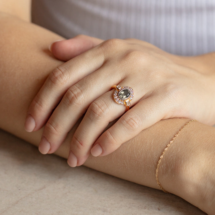 A close-up of hands on a table, featuring the Oval Deco Green Amethyst and CZ Ring with a large green amethyst surrounded by clear stones, paired with a delicate 14k gold vermeil chain bracelet on the wrist.