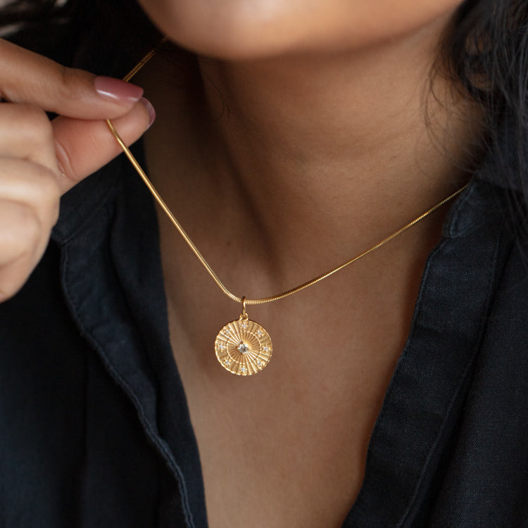 A person in a black top holds the Charms necklace by Carrie Elizabeth Jewelry, featuring a gold sunburst pendant with a small central gem and intricate texture, inspired by statement charms.