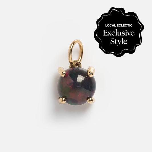 A round, dark One of a Kind 14k Small Black Welo Opal Charm pendant features subtle red and green hues in a yellow gold prong setting with a small loop. A “Local Eclectic Exclusive Style” badge highlights its unique opal jewelry appeal.