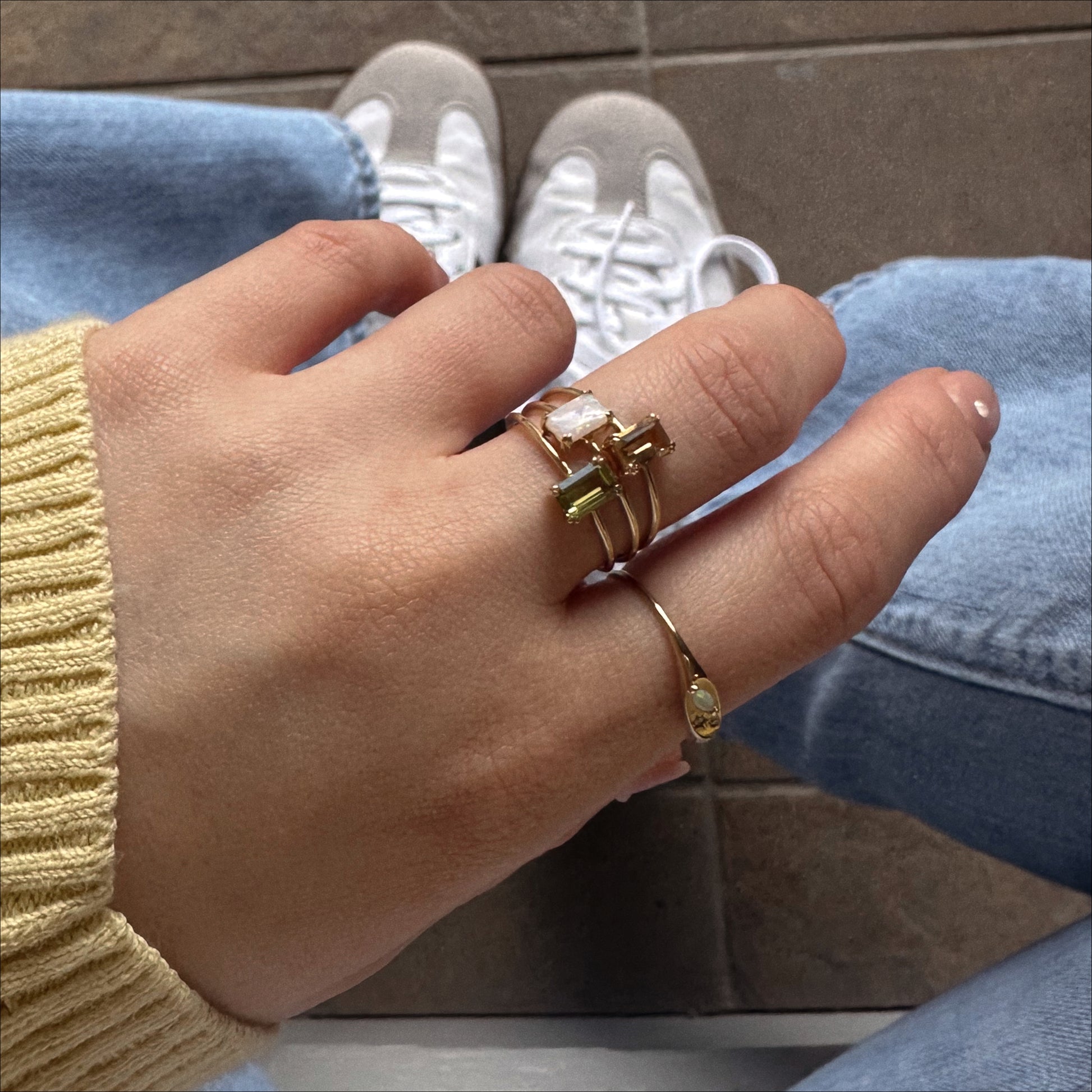 A hand wearing three gold rings, including the Solid Gold Essential Baguette Birthstone Ring and one with a cross design, rests on a knee. The person is dressed in a yellow sweater, blue jeans, and white sneakers.
